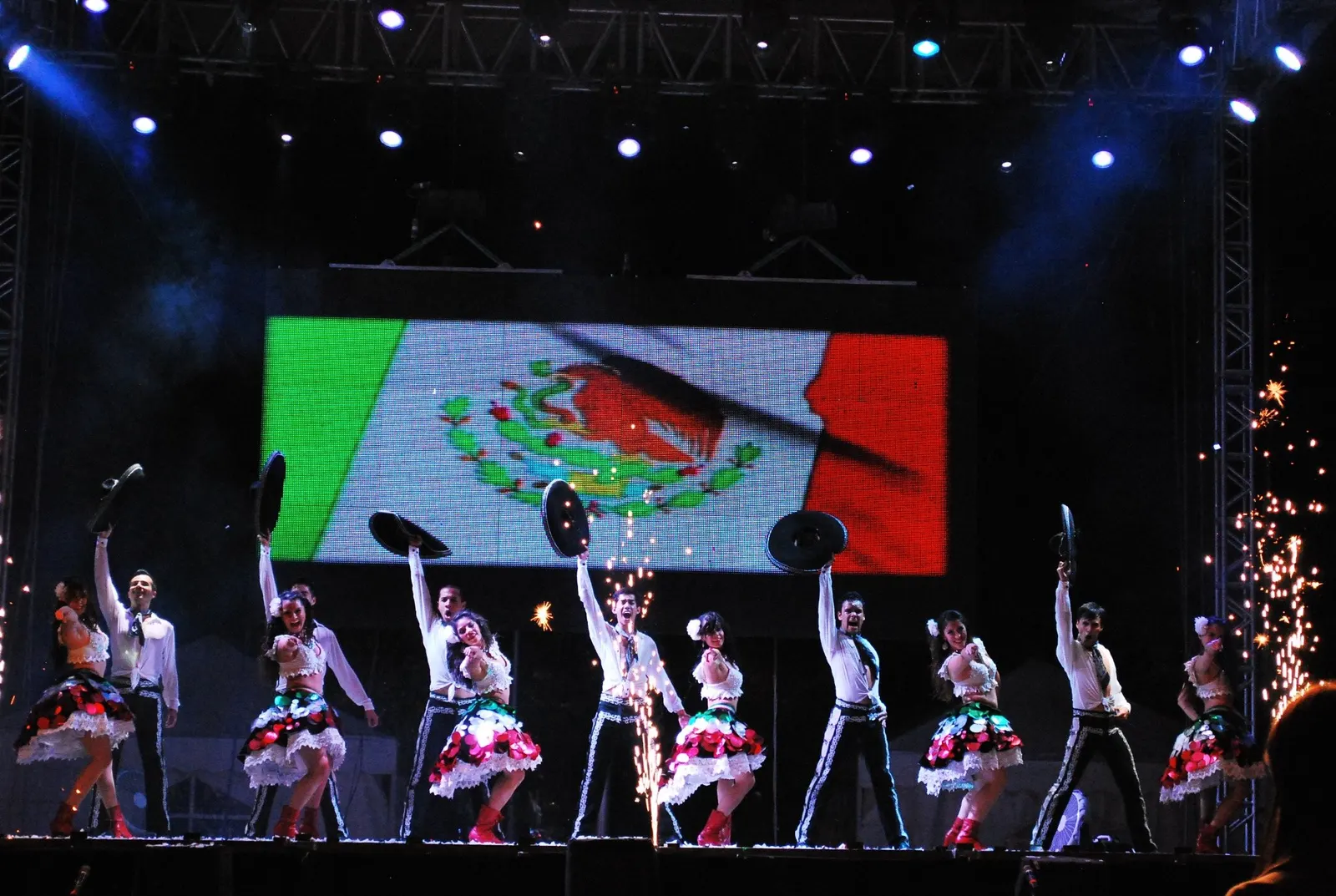 Espectáculo de danza mexicana en un escenario con la bandera de México, iluminación y efectos de pirotecnia